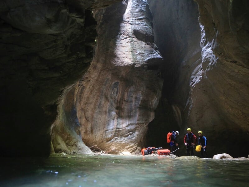 Groupe de personnes en combinaisons et casques dans un canyon avec de hautes parois rocheuses éclairées par le soleil.
