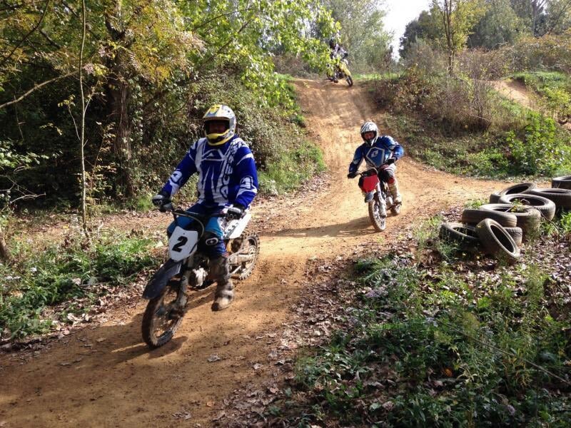 Deux pilotes de motocross en pleine course sur un sentier en forêt, avec des pneus usagés empilés en bordure et des collines terreuses en arrière-plan