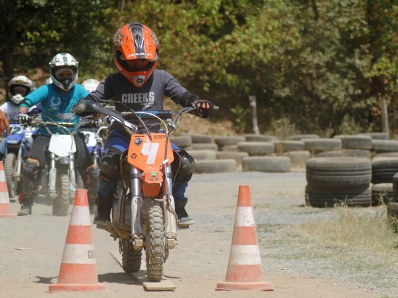 Enfant en motocross, portant un casque orange et une tenue sombre, franchissant un slalom de cônes sur un terrain poussiéreux, suivi par d'autres participants sur de petites motos