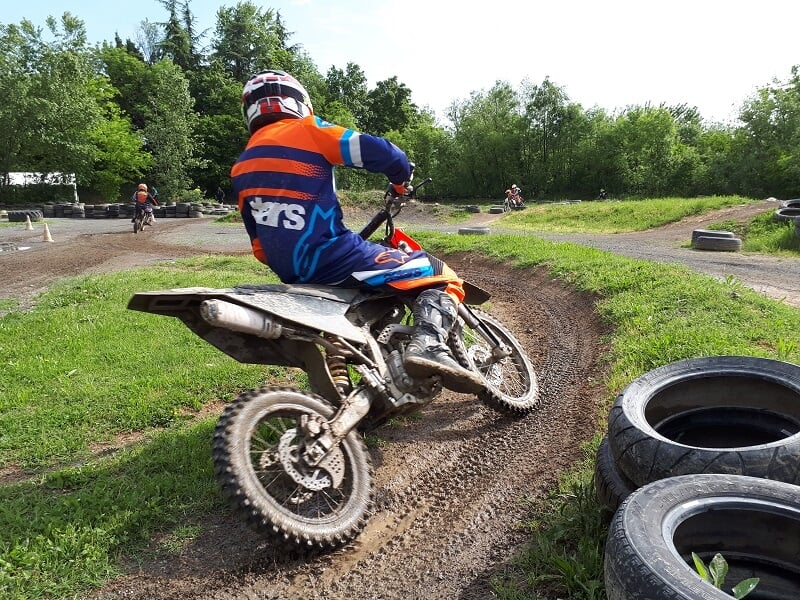 Motard en combinaison orange et bleue abordant un virage sur une piste en terre battue, avec des pneus disposés en bordure pour délimiter le tracé, et des arbres en arrière-plan