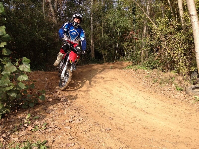 Pilote de motocross en tenue de protection bleue et rouge prenant un virage sur un chemin de terre entouré de forêt dense, soulevant de la poussière derrière lui