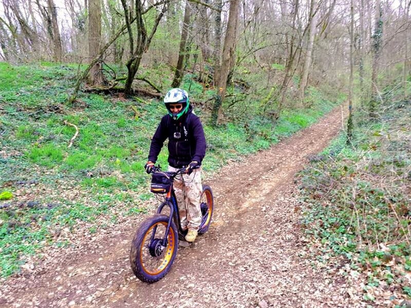 Individu en casque noir et vêtements sombres roulant sur une trottinette électrique dans une forêt.