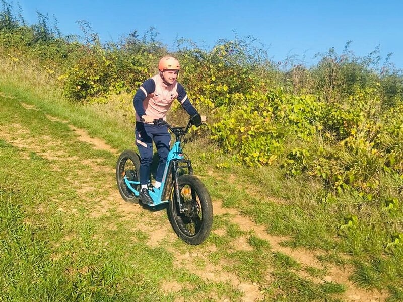 Homme souriant en casque orange conduisant une trottinette électrique bleue sur un chemin ensoleillé.