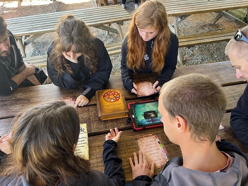 Plusieurs personnes concentrées autour d'une tablette et de documents, collaborant sur un jeu ou un puzzle à l'extérieur.