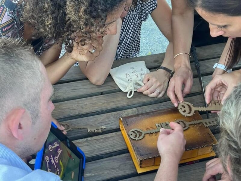 Groupe d'adultes manipulant des clés en bois et une boîte ornée, en plein air autour d'une table en bois.