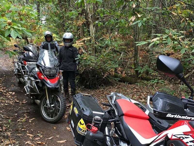 Deux motards en tenue complète posent à côté de leurs motos sur un chemin forestier en automne.