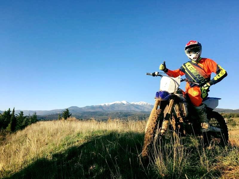 Motard en tenue orange et casque rouge pose avec sa moto tout-terrain sur une colline avec montagnes enneigées.