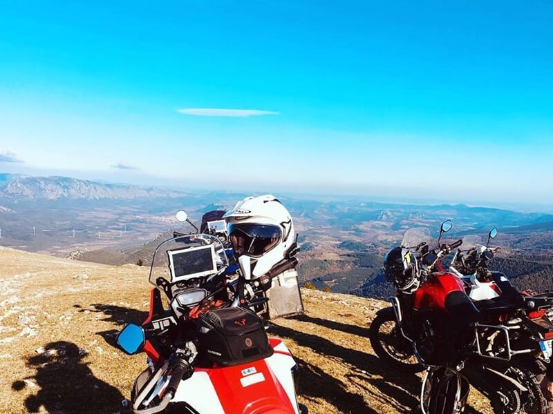 Deux motos stationnées en altitude avec vue panoramique sur une vallée montagneuse, ciel bleu dégagé, casque blanc posé sur le guidon.