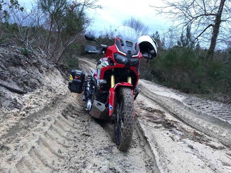 Moto tout-terrain rouge avec valises latérales sur un chemin sablonneux, entourée de buissons et d’arbres dénudés sous un ciel nuageux.