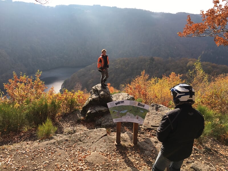 Deux personnes en tenue de moto regardent un panneau d'information sur un rocher avec un paysage de forêt en arrière-plan.
