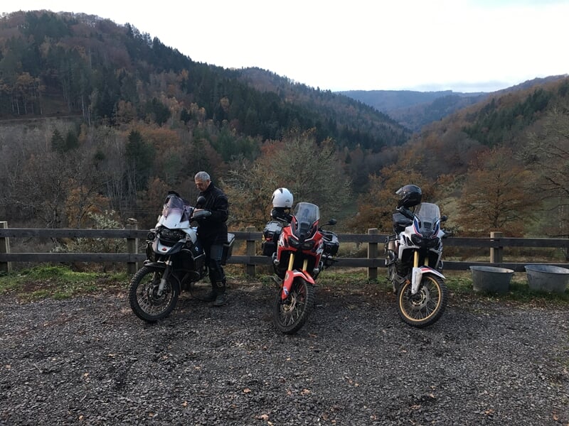 Trois motos garées sur un chemin de montagne avec des motards en tenue discutant dans un paysage automnal.