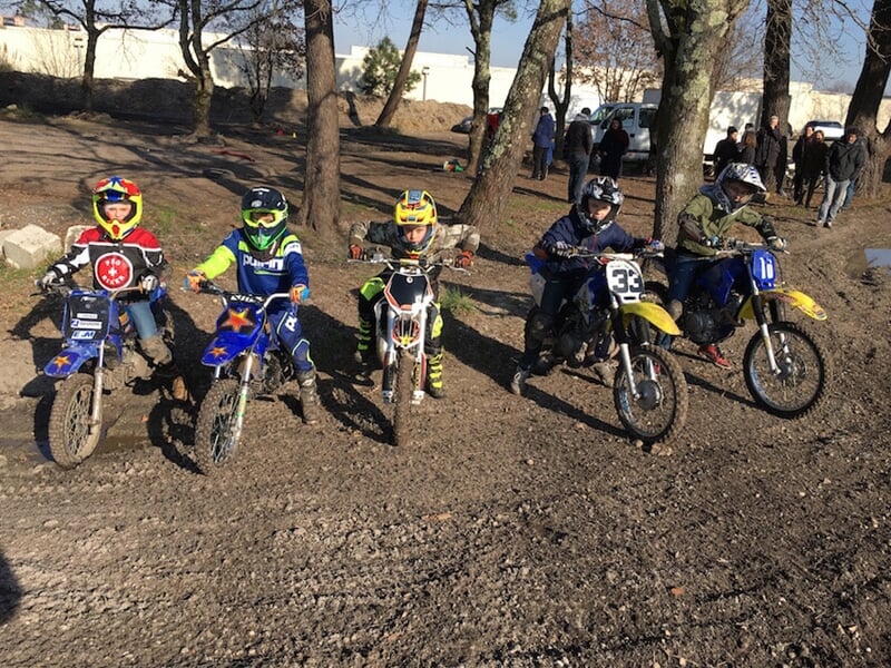 Cinq enfants en tenue de moto avec casques posent sur leurs motos sur un terrain de terre battue.