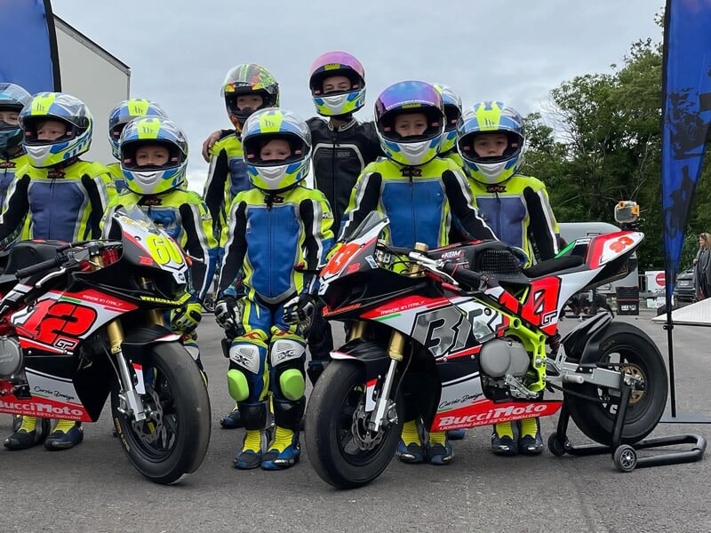 Groupe d'enfants en tenue de moto avec casques posent sur leurs motos sur un circuit asphalté en extérieur.