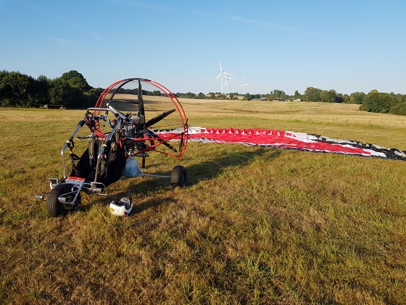 Paramoteur posé sur un champ herbeux avec voile rouge et blanche déployée, éoliennes en arrière-plan sous ciel bleu.