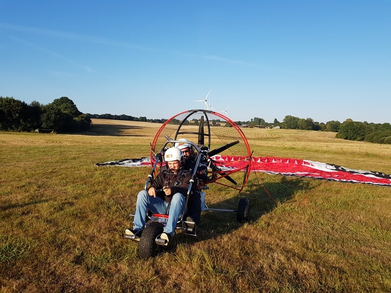 Personne assise dans un paramoteur au sol sur un champ, portant un casque blanc, voile rouge et blanche étalée derrière.