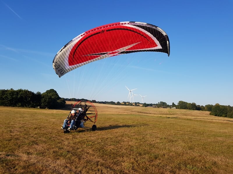 Paramoteur en train de décoller sur un champ, voile rouge et blanche gonflée, ciel bleu et éoliennes visibles au loin.