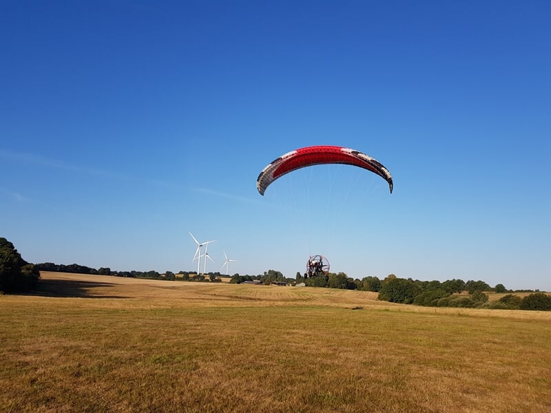 Paramoteur en vol au-dessus d'un champ, voile rouge et blanche déployée, ciel bleu clair et éoliennes en arrière-plan.