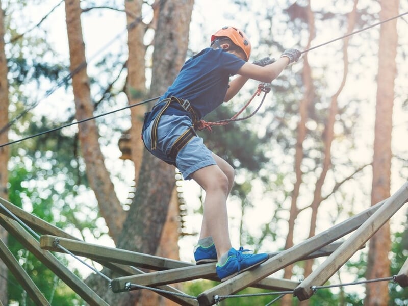 Enfant équipé d'un casque et harnais franchissant un pont suspendu en bois dans un parc accrobranche en forêt.