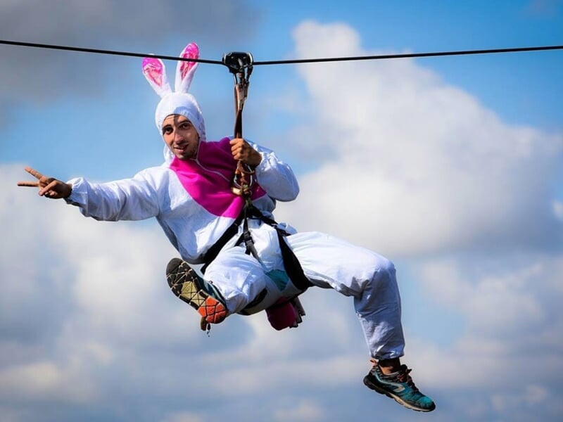 Personne déguisée en lapin blanc portant un casque et harnais, glissant sur une tyrolienne sous un ciel bleu.