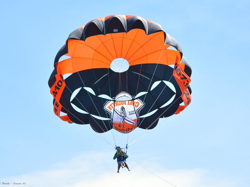 Parachute ascensionnel orange et noir avec une personne suspendue, ciel bleu clair en arrière-plan.