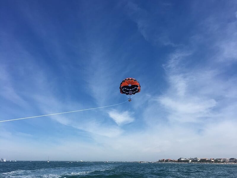 Parachute ascensionnel rouge et noir avec une personne suspendue, vue éloignée au-dessus de la mer et du ciel bleu clair.