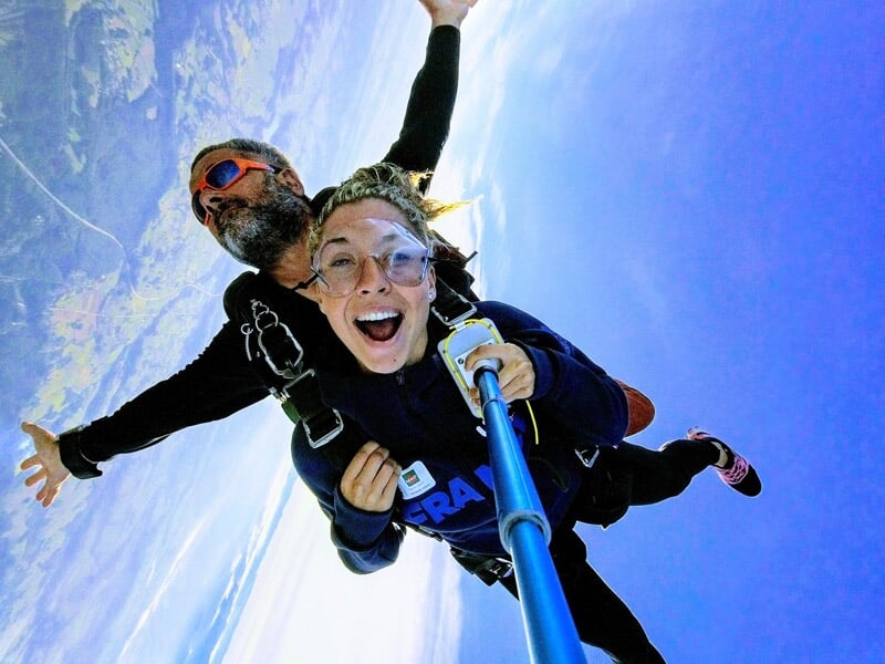 Duo de parachutistes en tandem capturant un selfie, large sourire et arrière-plan d’un paysage rural et côtier