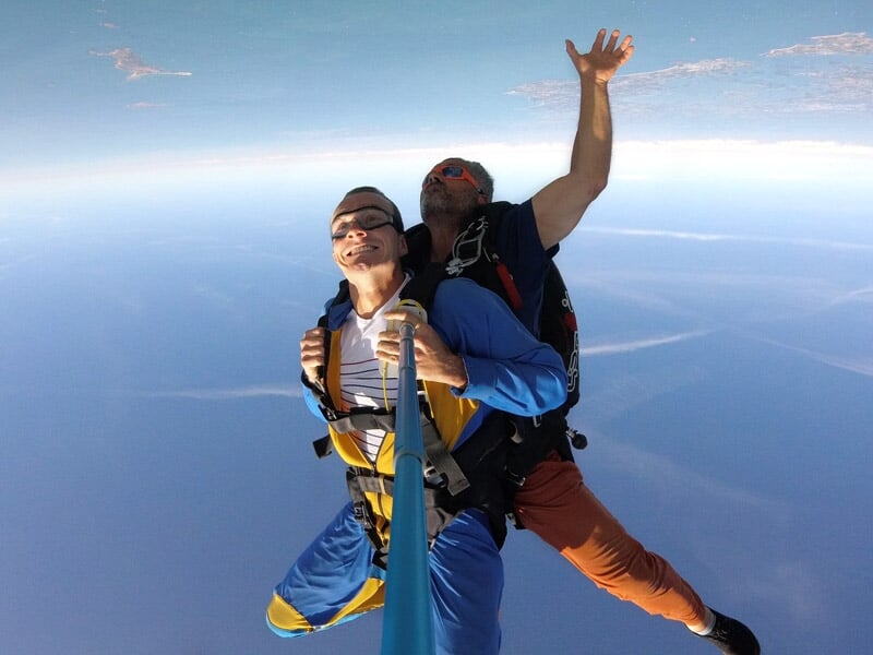 Parachutiste en tandem capturant un selfie en plein ciel, combinaison colorée, gestes enthousiastes, horizon dégagé et vue sur la mer en arrière-plan