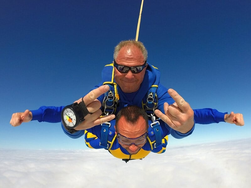 Parachutistes en tandem portant des combinaisons colorées, gestes festifs au-dessus des nuages sous un ciel bleu