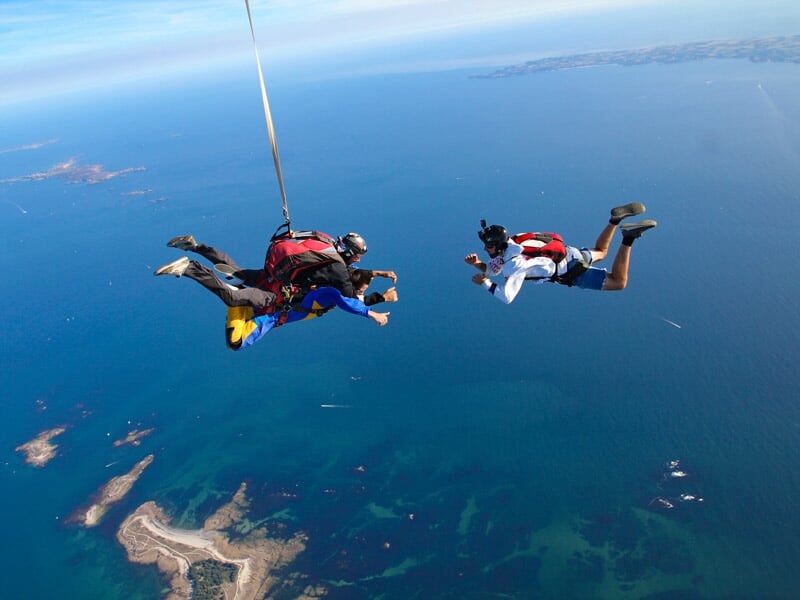 Saut en parachute tandem, deux parachutistes en chute libre au-dessus de l'océan, îles visibles en arrière-plan.