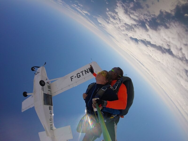 Saut en parachute tandem, homme et instructeur en chute libre, avion visible en arrière-plan, ciel bleu avec nuages.