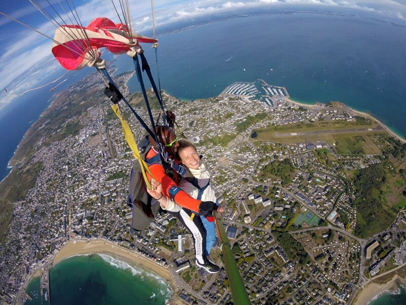 Saut en parachute tandem, femme souriante en combinaison rouge et blanche, parachute ouvert, vue aérienne d'une ville côtière et de la mer.