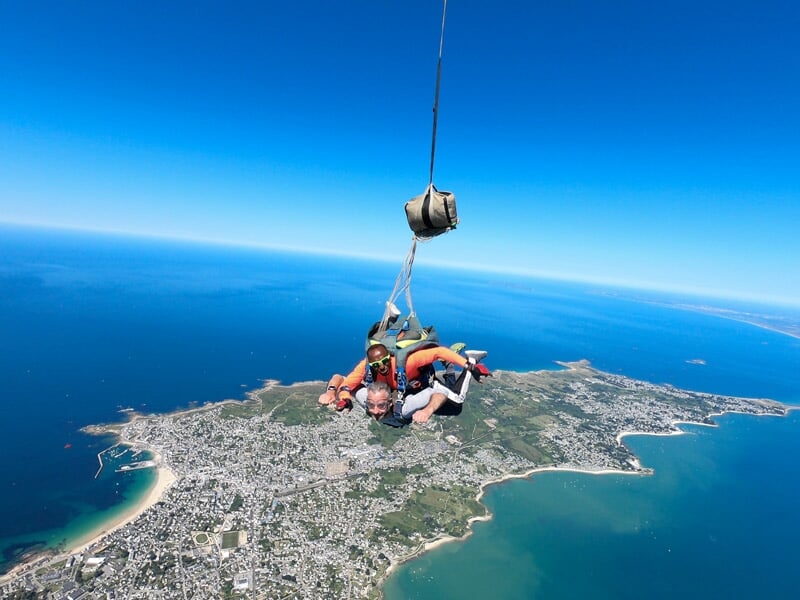 Saut en parachute tandem, homme et instructeur en chute libre, ville côtière et mer en arrière-plan, ciel bleu clair.