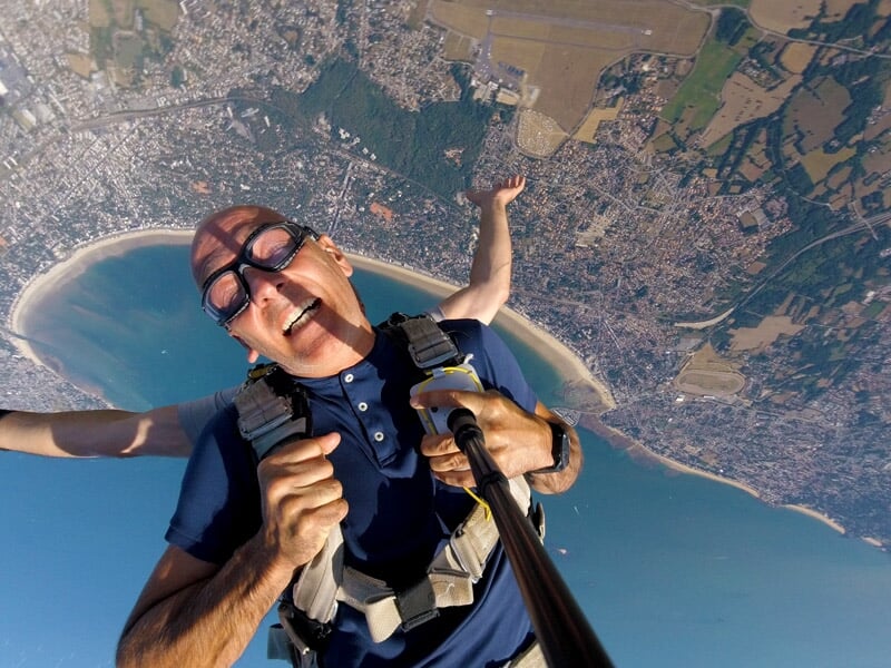 Parachutiste en tandem en plein vol, visage souriant, vue plongeante sur une baie et une ville côtière
