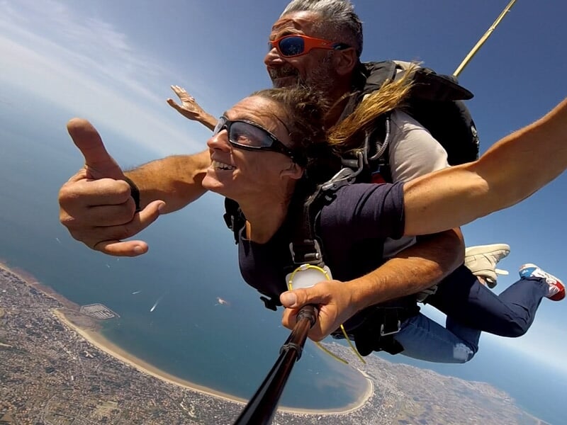 Saut en parachute en tandem avec gestes enthousiastes, mer et littoral visibles en contrebas, ciel dégagé