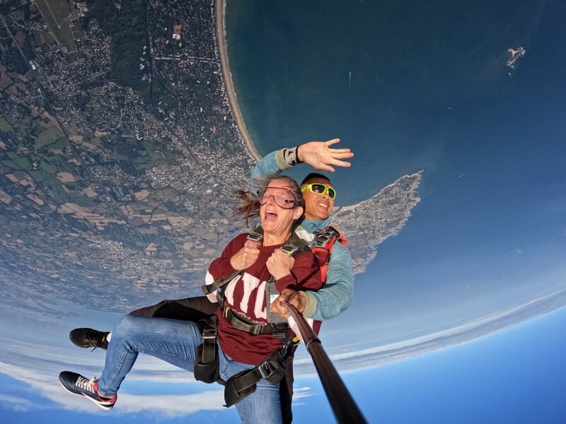 Parachutistes en tandem capturant leur saut en selfie, expressions joyeuses, panorama côtier avec plage et campagne en arrière-plan