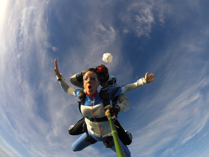 Parachutiste en tandem capturé en plein vol, expressions d'excitation et ciel bleu parsemé de nuages, parachute stabilisateur visible