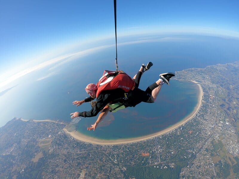 Saut en parachute tandem, homme en combinaison rouge et noire, parachute ouvert, vue aérienne d'une grande plage courbée et d'une ville côtière, mer en arrière-plan.