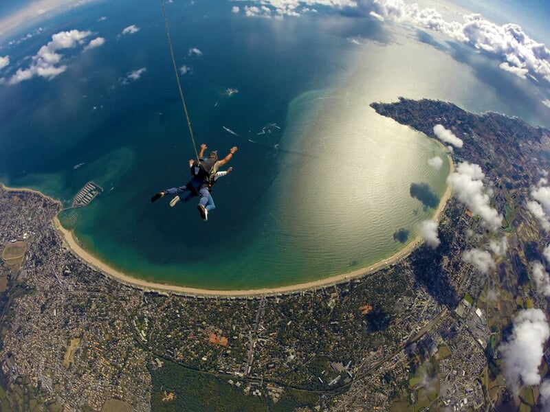 Saut en parachute au-dessus d'une grande baie ensoleillée avec une plage en arc de cercle et une ville en contrebas, ciel dégagé avec quelques nuages légers