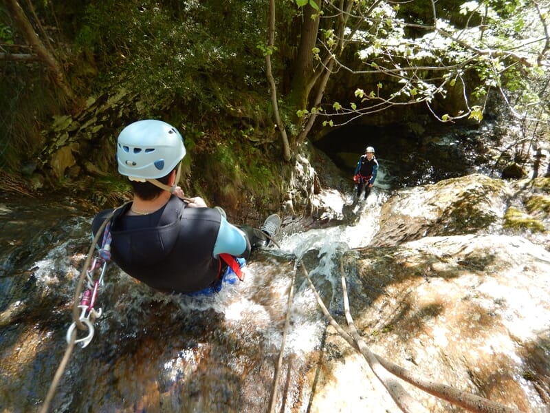 Personne en casque bleu descend une cascade rocheuse en canyoning dans un environnement naturel verdoyant.