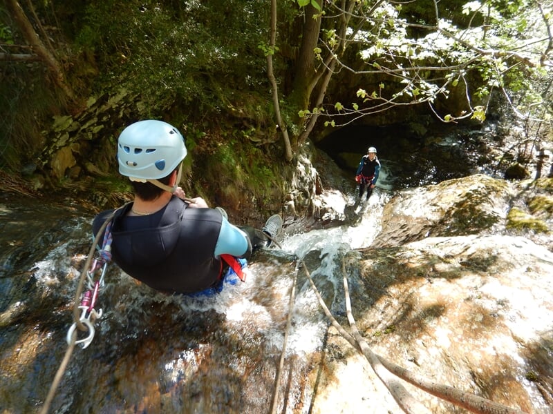 Personne en casque bleu descend une cascade rocheuse en canyoning dans un environnement naturel verdoyant.