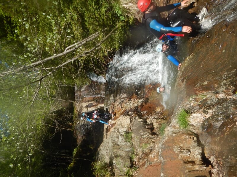 Groupe de personnes en combinaisons et casques rouges et bleus glissent sur des rochers mouillés en canyoning.