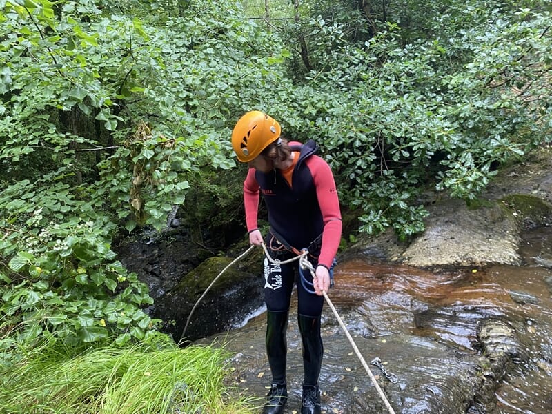 Femme en casque orange et combinaison noire prépare une descente en rappel au bord d'un cours d'eau.