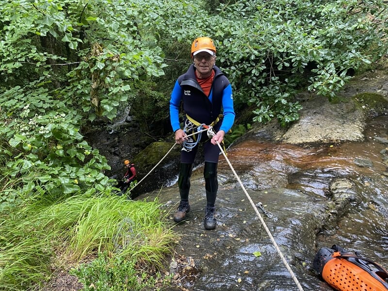 Homme en casque orange et combinaison bleue tient une corde pour une activité de canyoning en forêt.
