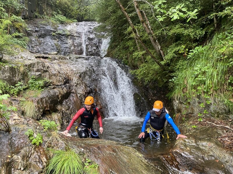Deux personnes en combinaisons et casques oranges progressent dans un ruisseau avec une cascade en arrière-plan.