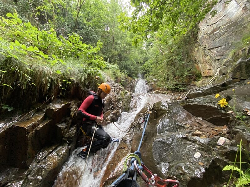 Personne en casque orange et combinaison noire escalade une cascade rocheuse équipée de cordes en canyoning.