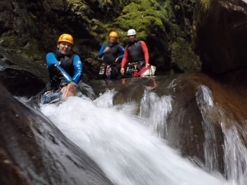 Trois personnes en casque et combinaison de canyoning glissent sur un toboggan naturel dans une rivière.