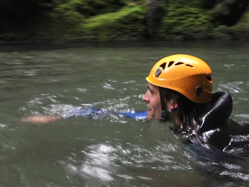 Un homme en casque orange nage dans une rivière calme entourée de végétation dense.