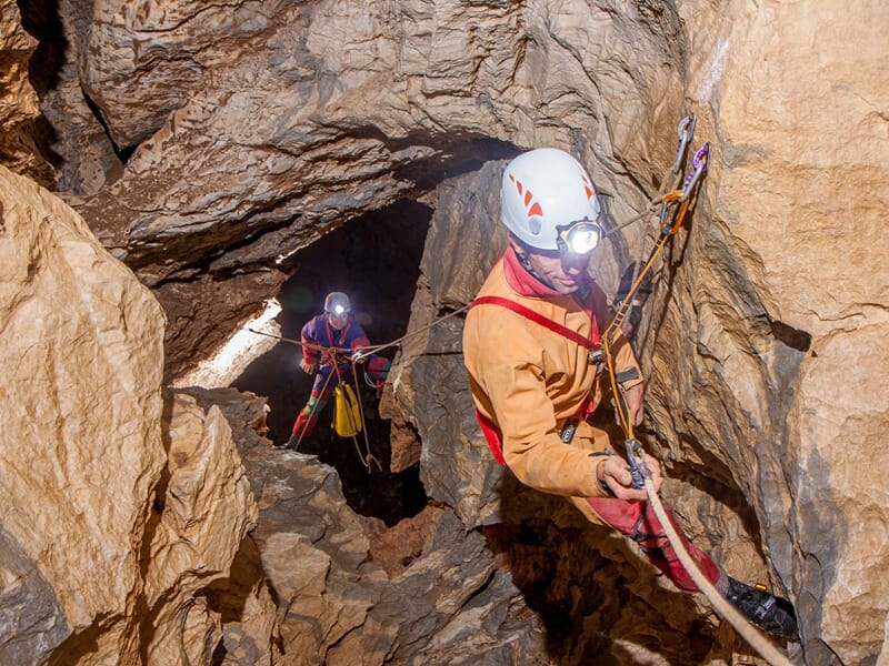 Deux spéléologues équipés de casques et lampes frontales descendent dans une grotte rocheuse étroite et sombre.