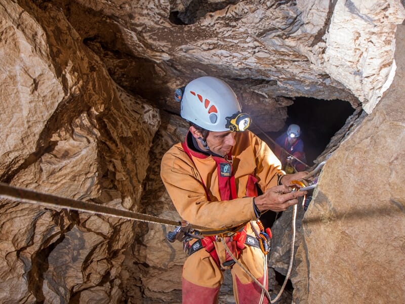 Spéléologue en combinaison orange vérifiant son équipement d'escalade dans une cavité rocheuse avec un autre en arrière-plan.