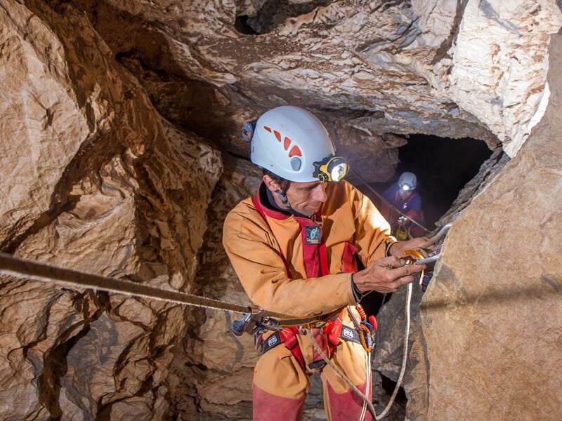 Spéléologue en combinaison orange vérifiant son équipement d'escalade dans une cavité rocheuse avec un autre en arrière-plan.
